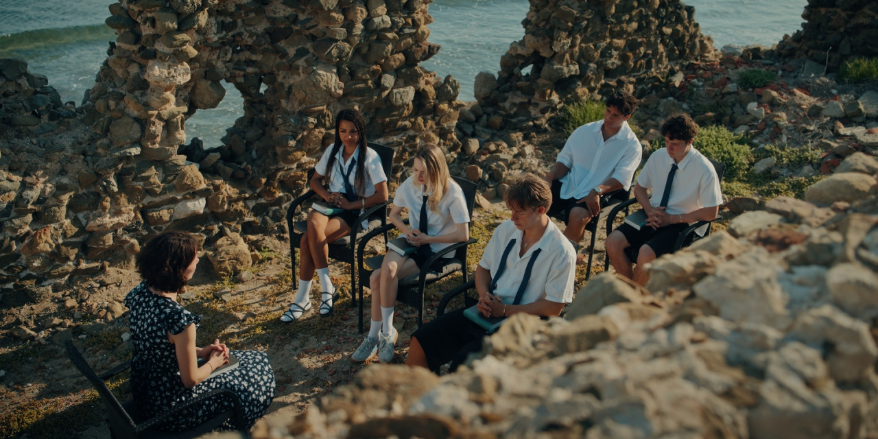 Gruppe Schüler sitzt zur Bibelstunde am Strand. Szenenbild aus Prime Video Serie 'Die neue Generation Krass Klassenfahrt'.
