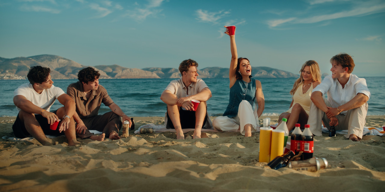 Eine Gruppe Jugendlicher am Sandstrand bei warmem Tageslicht. Frame aus Prime Video Serie 'Die neue Generation Krass Klassenfahrt'.